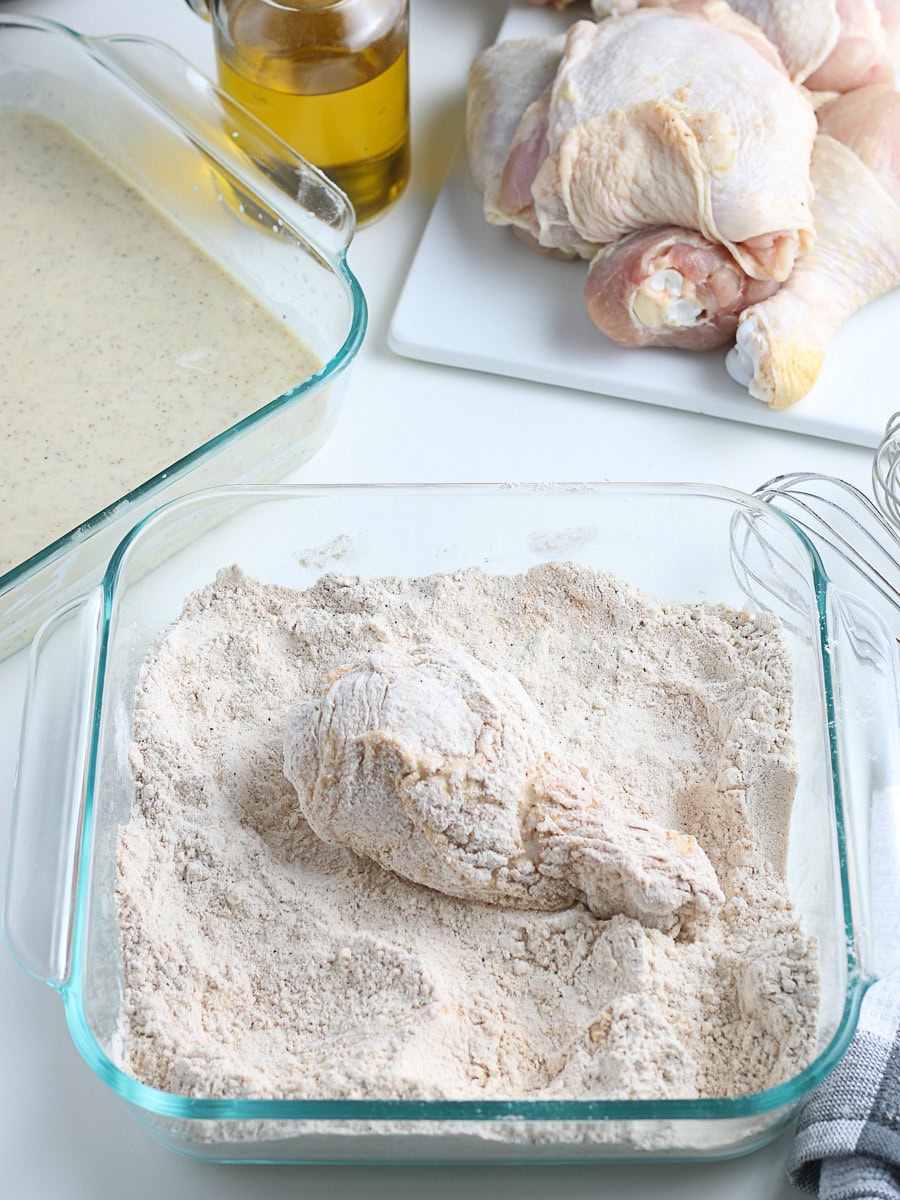 Raw chicken drumstick coated in flour sits in a glass dish, with a tray of flour, marinated chicken pieces, and a bottle of oil visible in the background.