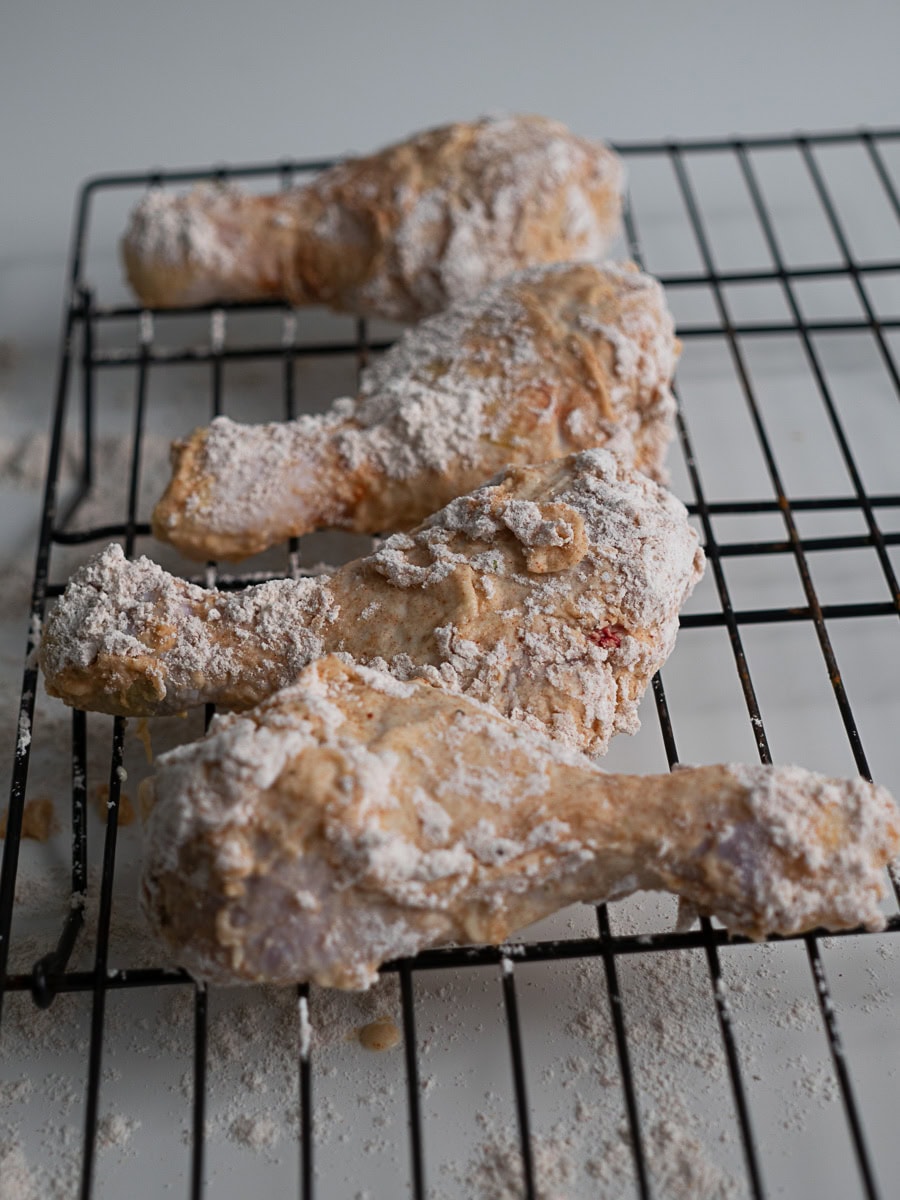 Four uncooked, flour-coated chicken drumsticks rest on a black wire cooling rack over a white surface.