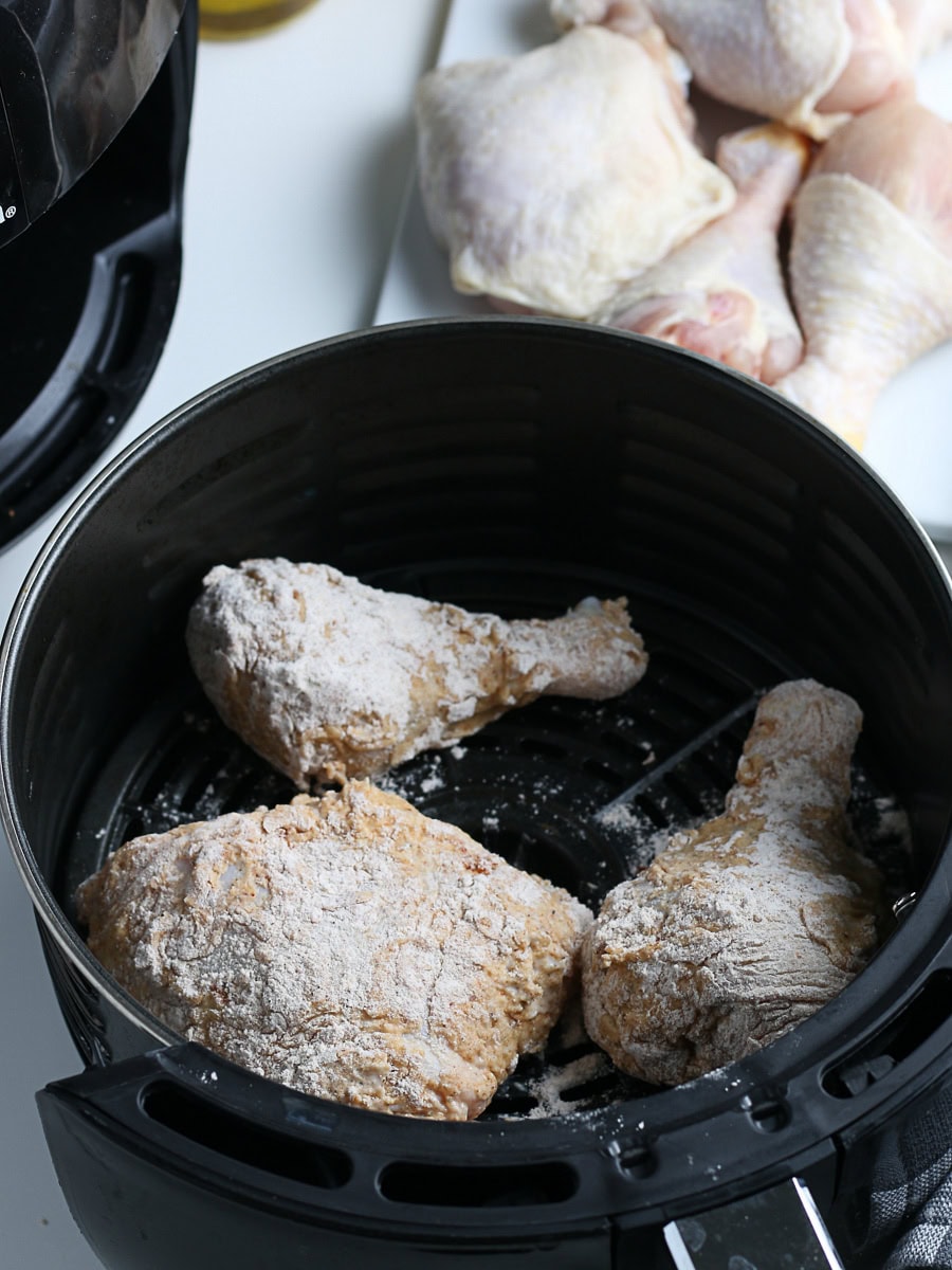 Three breaded chicken drumsticks are placed inside an air fryer basket, with raw chicken pieces on a plate in the background.
