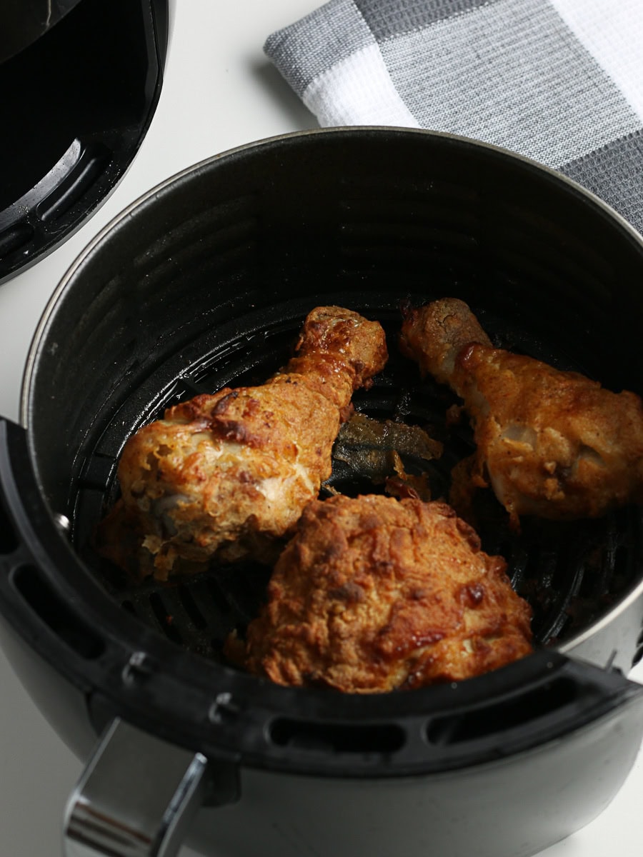 Three pieces of cooked fried chicken are in the basket of an air fryer, with a folded kitchen towel nearby on the white countertop.