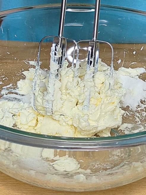 Electric beaters mixing butter and sugar in a glass bowl on a wooden surface.