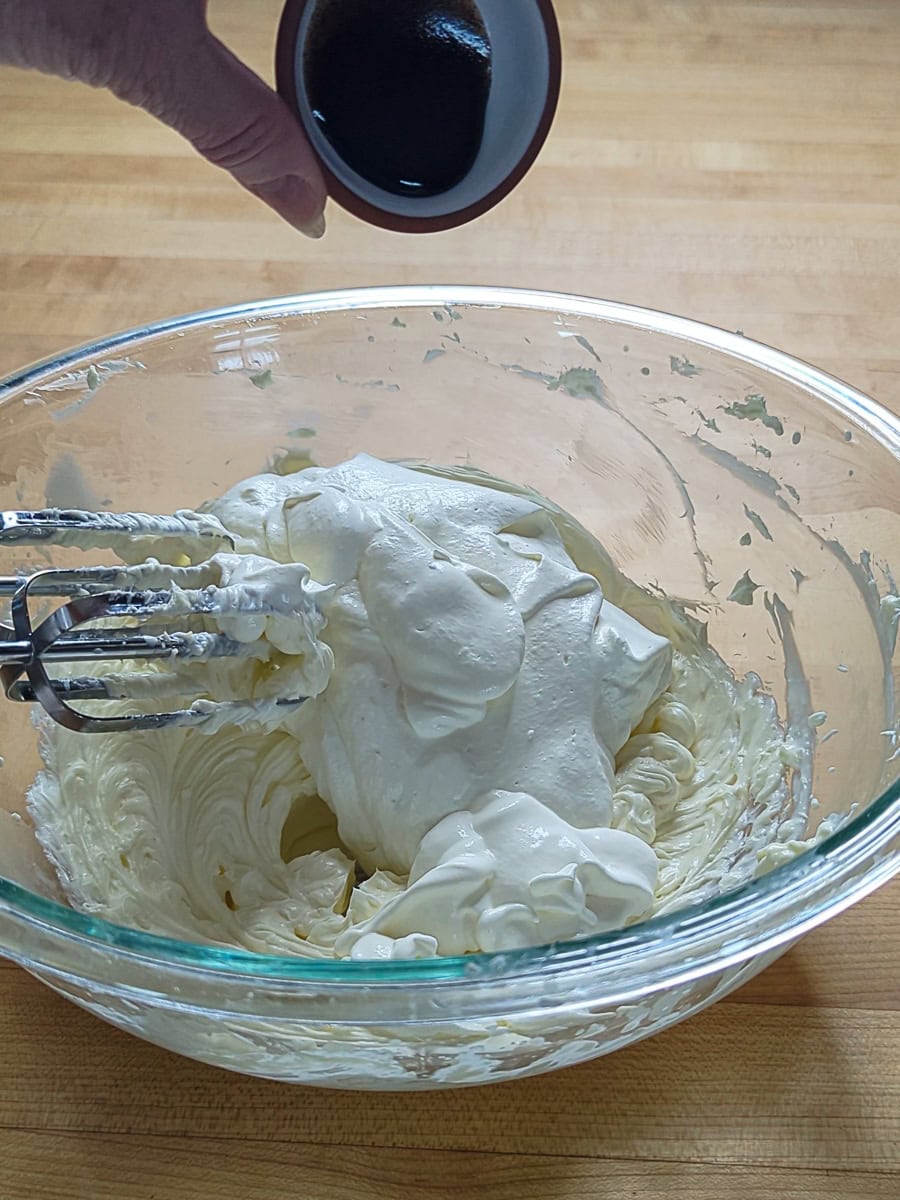 A hand holds a small bowl of dark liquid above a glass mixing bowl containing whipped cream and an electric beater on a wooden countertop.