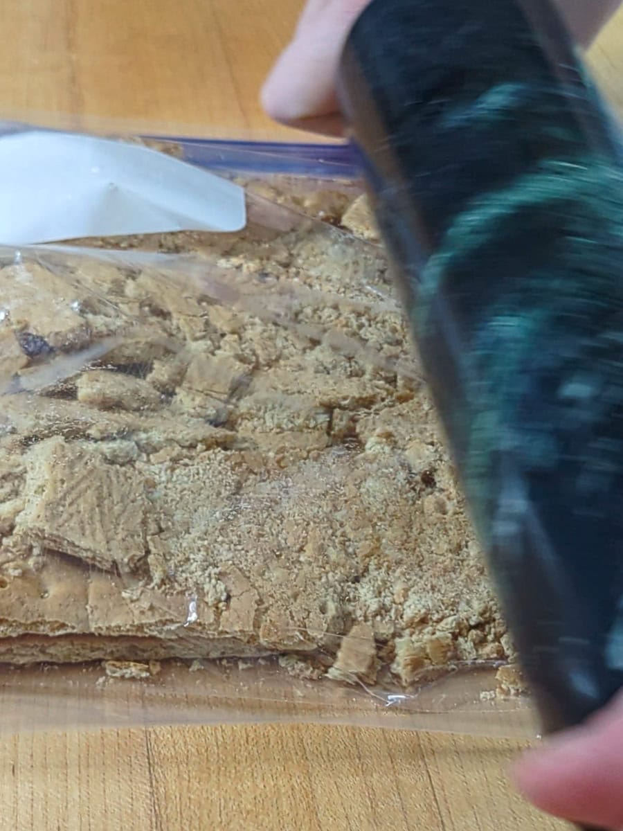 A person uses a rolling pin to crush graham crackers inside a plastic bag on a wooden surface.