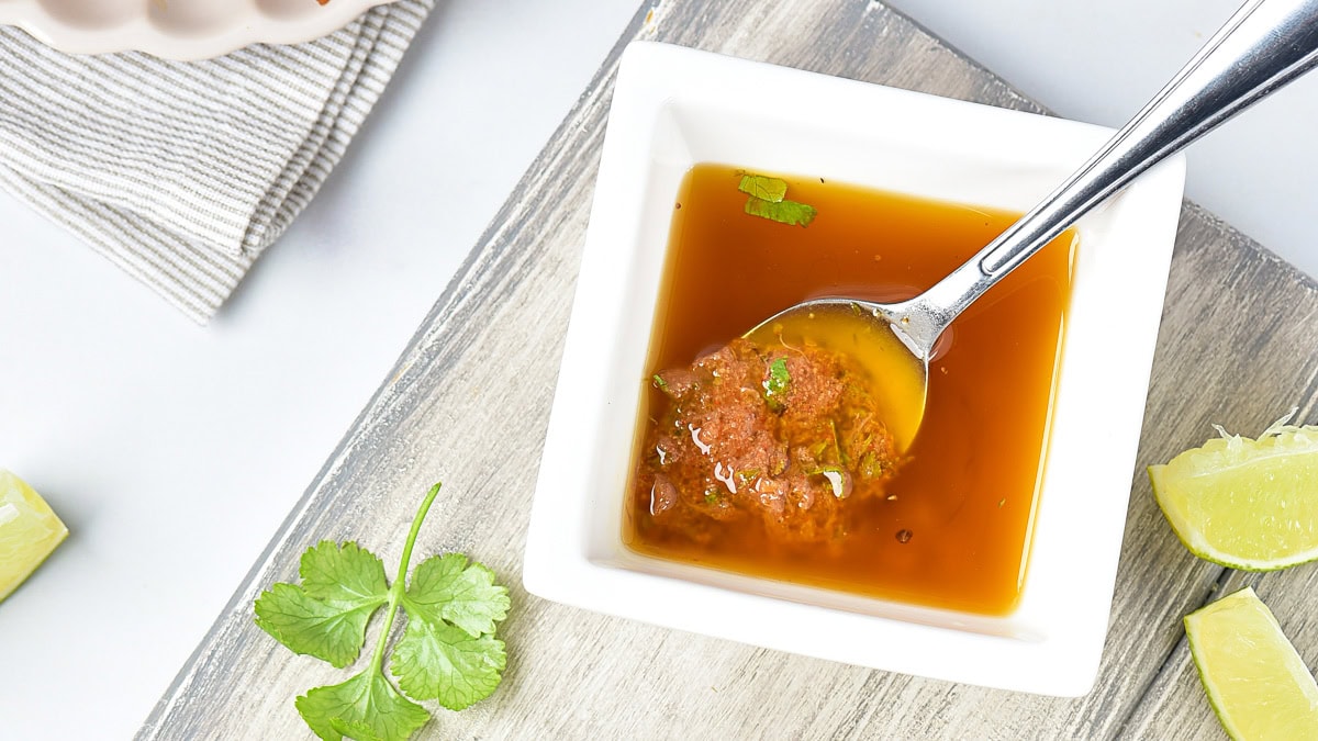 A square white bowl with brown sauce and herbs, a metal spoon inside, lime wedges, and a cilantro leaf on a wooden surface nearby.