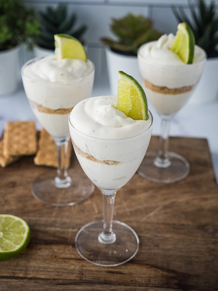 Three glasses of key lime mousse with graham cracker crumbs, each topped with a lime wedge, are displayed on a wooden surface with graham crackers and a lime nearby.