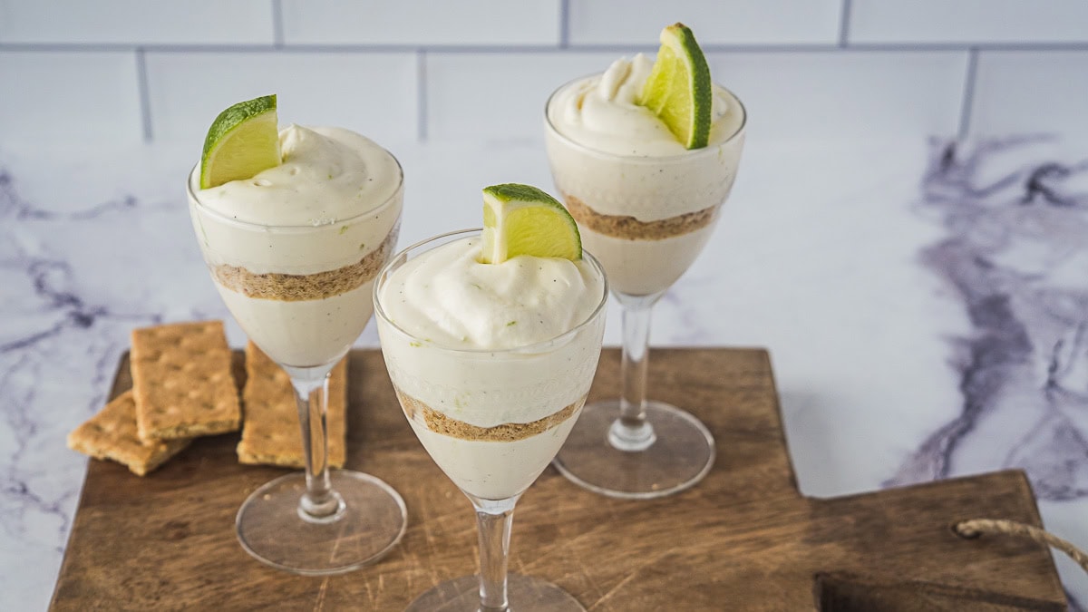 Three layered dessert parfaits in wine glasses, garnished with lime wedges, are displayed on a wooden board with graham crackers in the background.