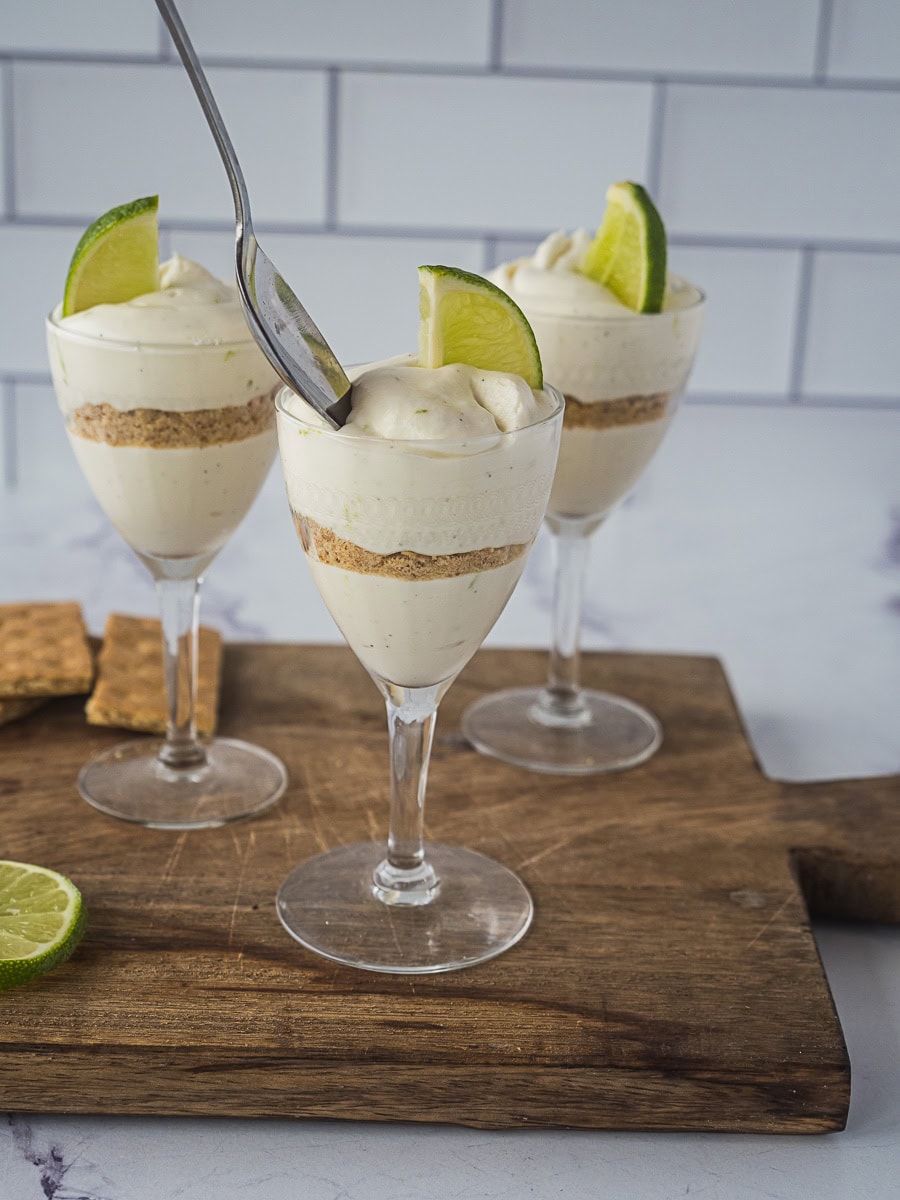 Three parfait glasses filled with layered cheesecake mousse and graham cracker crumbs, each topped with a lime wedge, are displayed on a wooden board; one glass has a spoon in it.