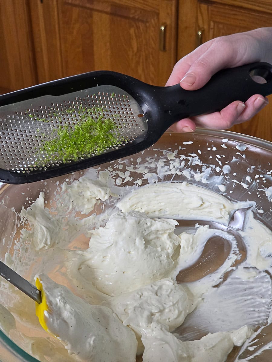 A hand holds a grater with lime zest over a glass bowl containing creamy white mixture, with a spatula resting inside the bowl.