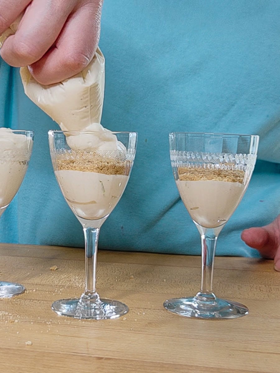 A person pipes creamy dessert mixture into a wine glass layered with crumbs, with two similar glasses beside it on a wooden surface.