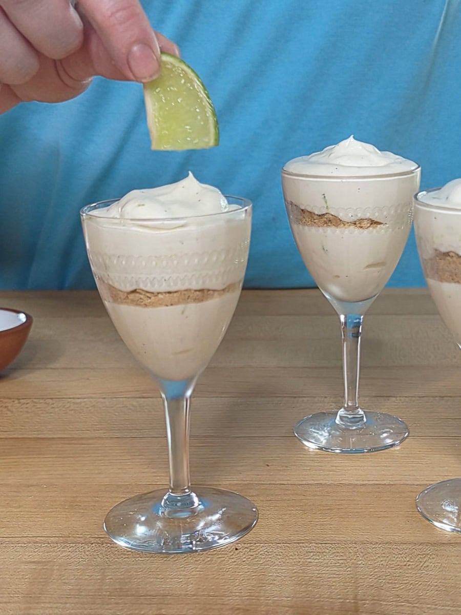 A hand holds a lime wedge over a dessert in a glass, layered with cream and crumbs, on a wooden surface with two similar desserts in the background.