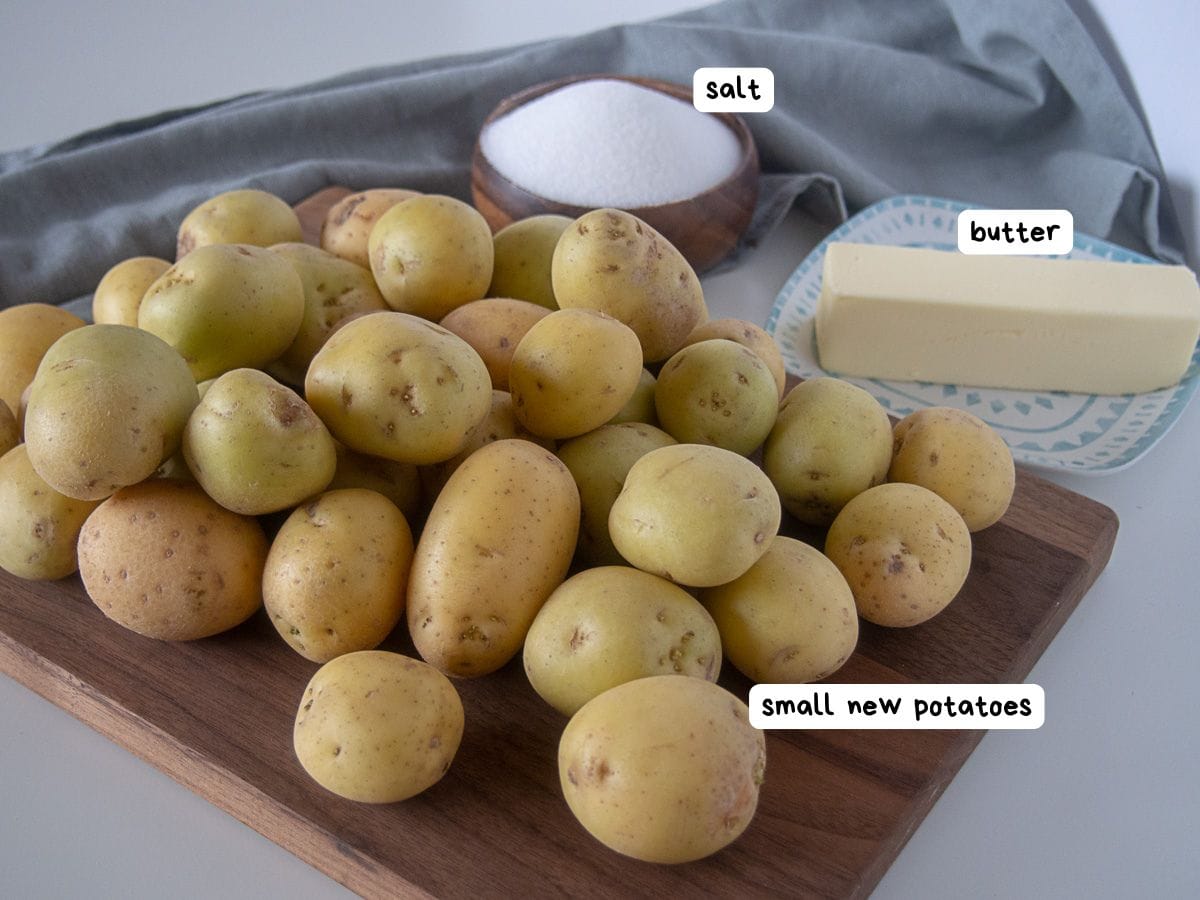 A pile of small new potatoes on a wooden board, with a stick of butter and a bowl of salt in the background.