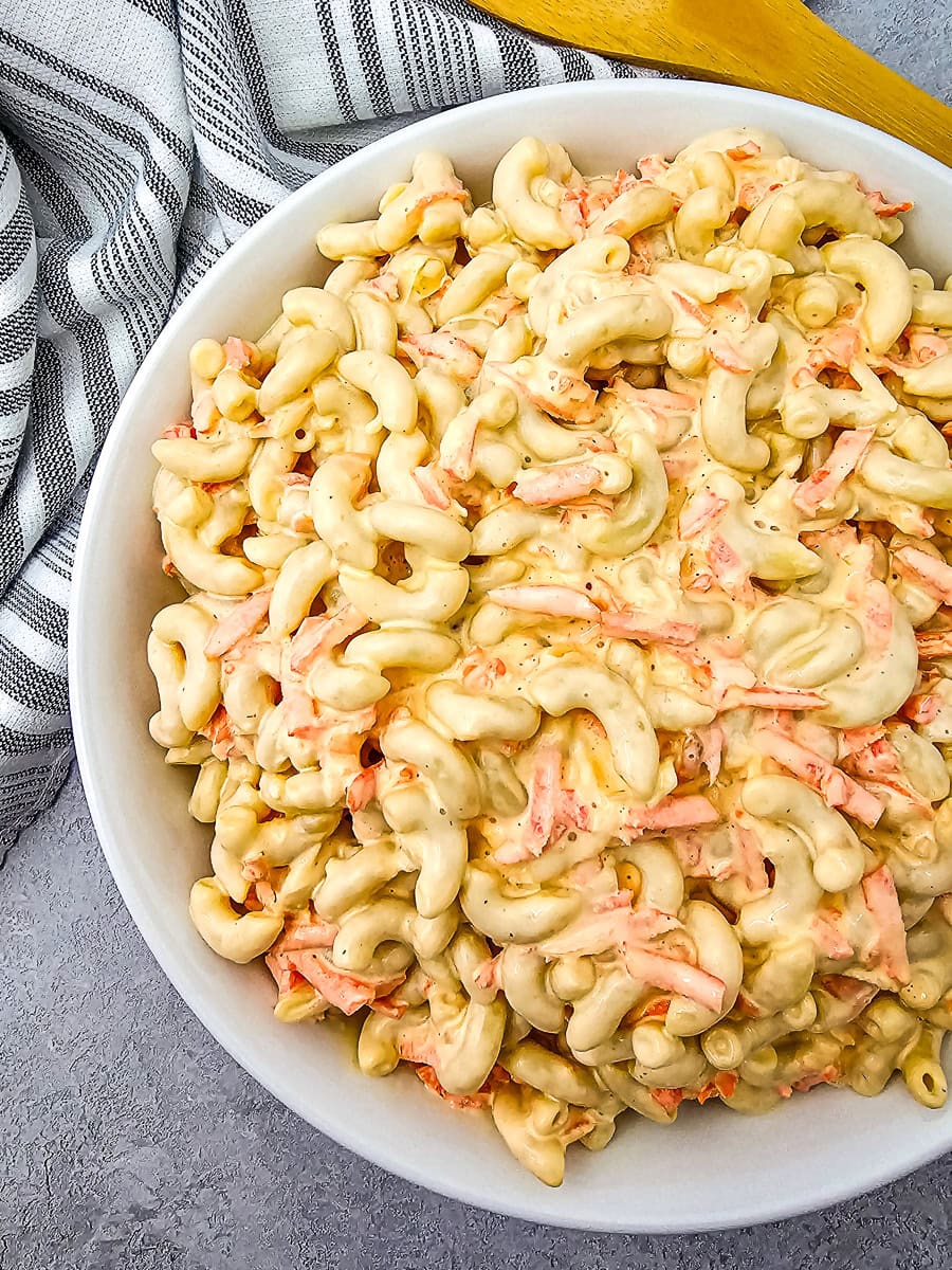 A bowl of macaroni salad with elbow pasta and shredded carrots in a creamy dressing, placed on a gray surface next to a striped cloth and a wooden spoon.