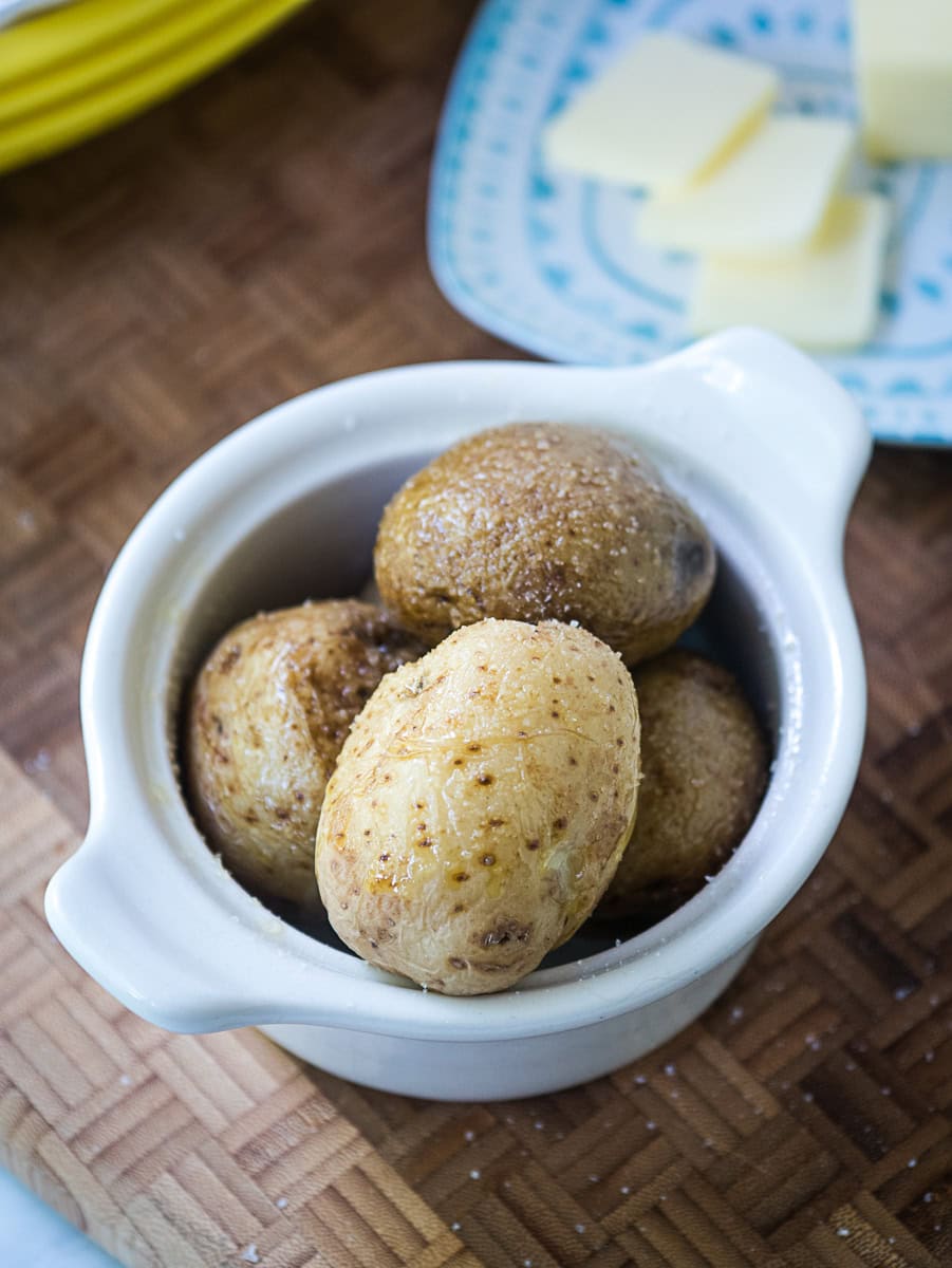 Small baked potatoes sprinkled with salt in a white ceramic dish, with a plate of sliced butter in the background.