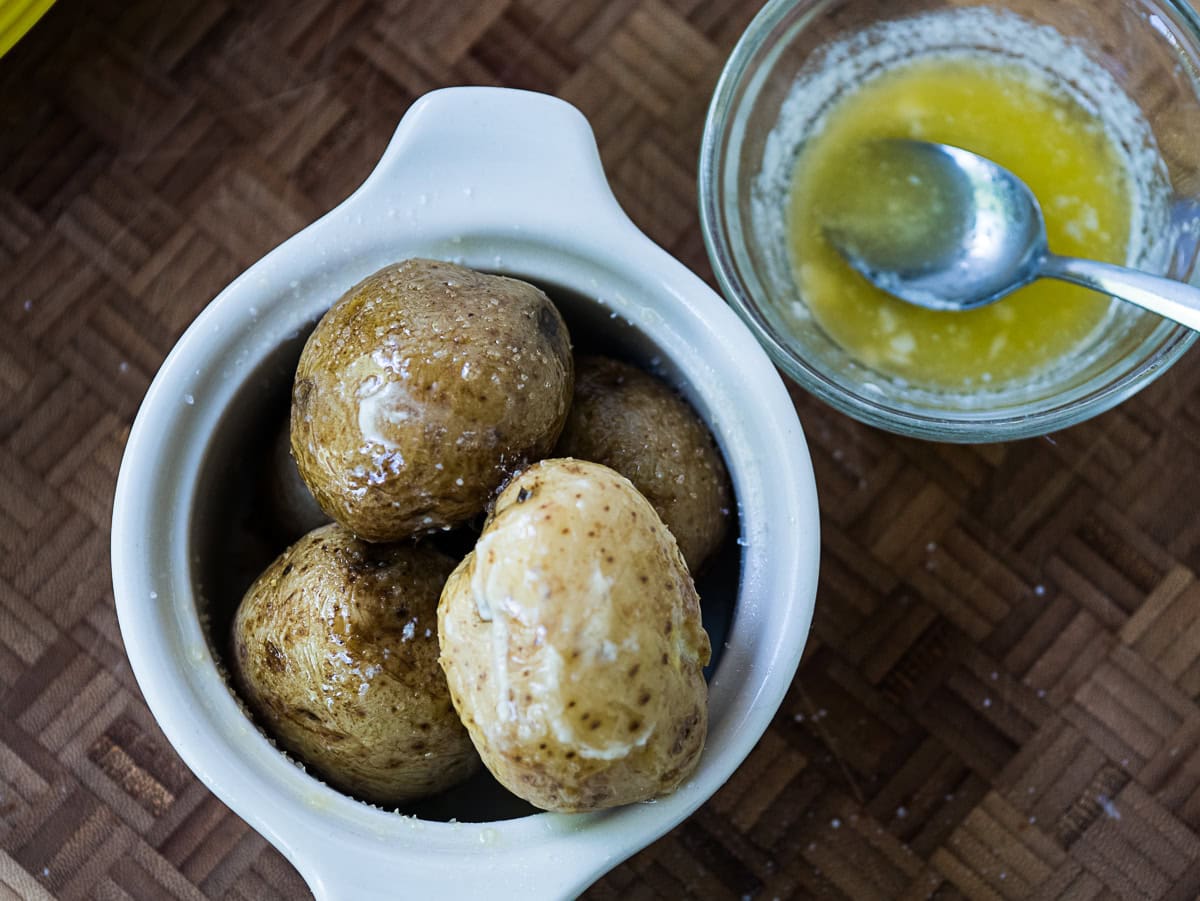 A small white dish with four boiled potatoes next to a glass bowl containing melted butter and a metal spoon on a wooden surface.