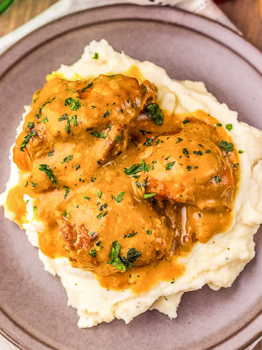 Three pieces of chicken with brown gravy and chopped herbs served on a bed of mashed potatoes on a plate.