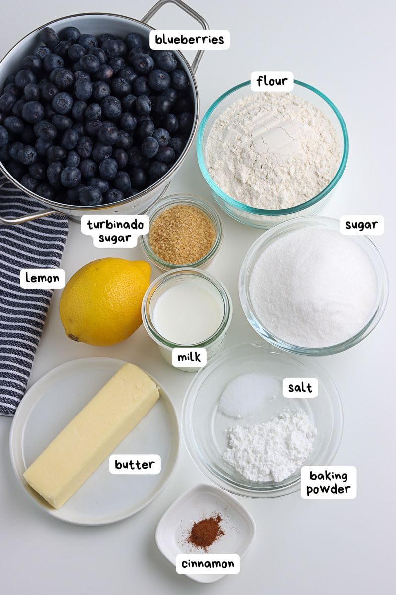 Assorted baking ingredients for blueberry cobbler, including blueberries, flour, sugar, turbinado sugar, lemon, milk, butter, salt, baking powder, and cinnamon arranged on a white surface.