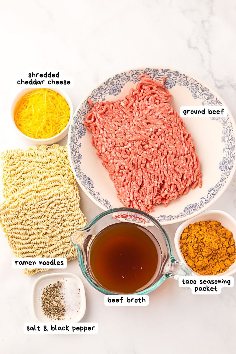 A plate of raw ground beef, shredded cheddar cheese, uncooked ramen noodles, beef broth, taco seasoning packet, and a small dish of salt and black pepper on a white surface.