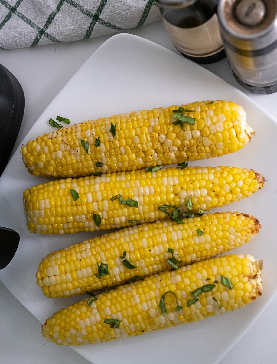 Four ears of cooked corn on the cob garnished with chopped herbs sit on a white square plate, with salt and pepper shakers nearby.