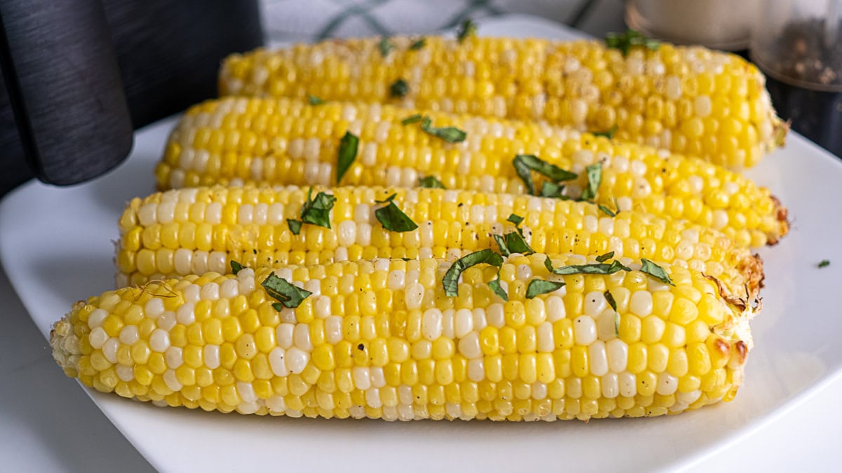 Four ears of cooked corn on the cob, garnished with chopped herbs, arranged on a white plate.