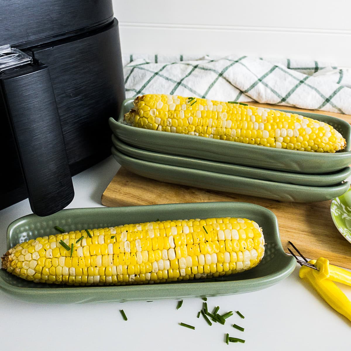 Two ears of cooked corn on the cob topped with chopped chives are served on green plates next to an air fryer, a corn peeler, and a checkered towel.