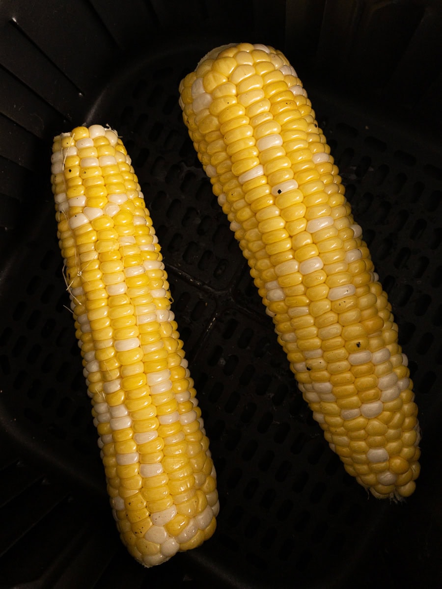 Two ears of yellow and white corn are placed side by side inside a black air fryer basket.