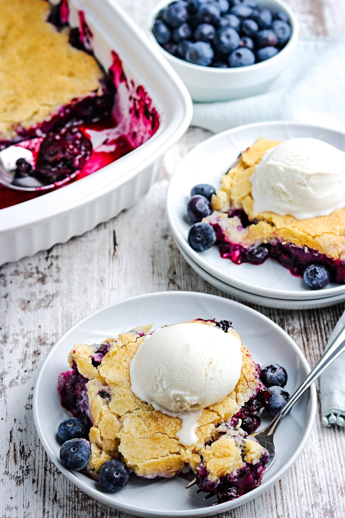Two plates of blueberry cobbler topped with vanilla ice cream and fresh blueberries, with a baking dish of blueberry cobbler and a bowl of blueberries in the background.