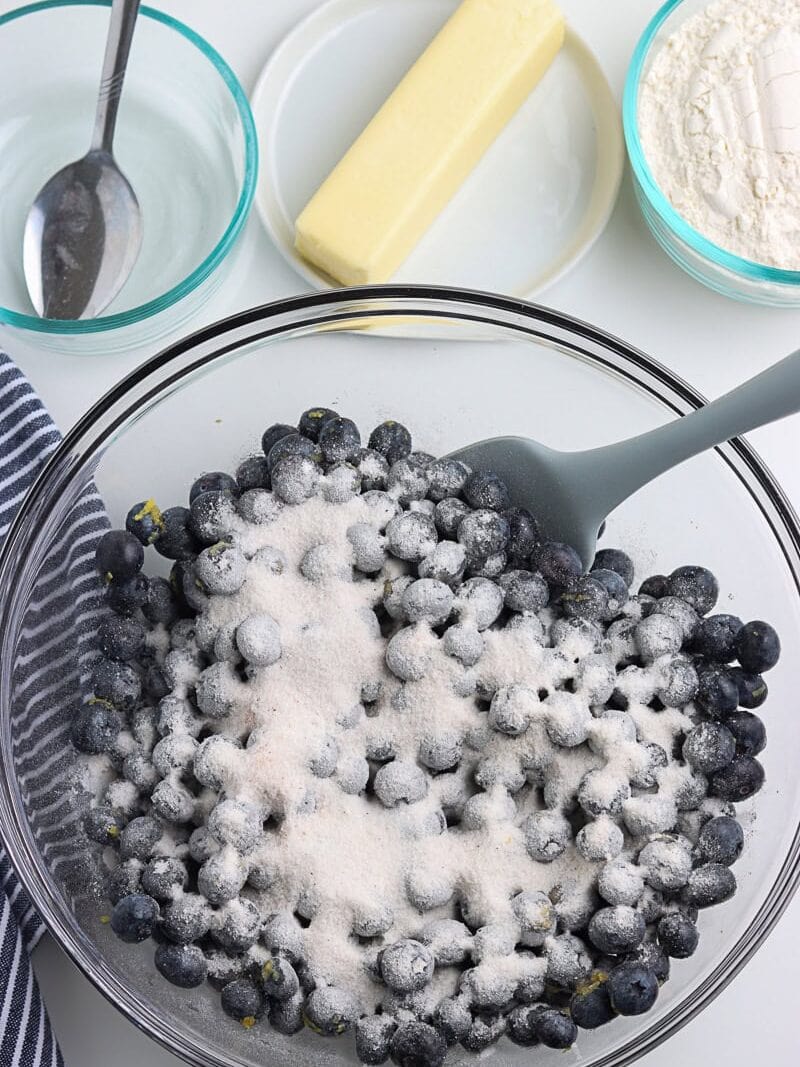 A glass bowl of blueberries coated with sugar, next to a mixing spoon, with bowls of flour, a stick of butter, and a spoon with liquid nearby.