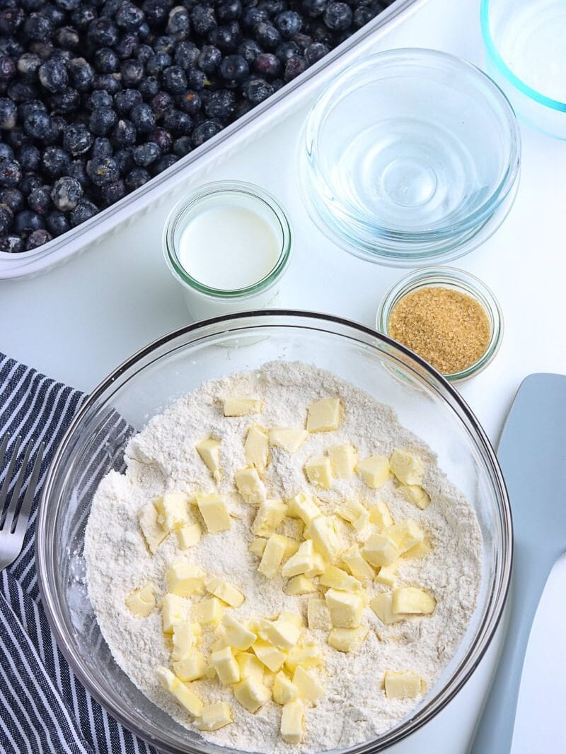A bowl of flour with cubed butter, surrounded by bowls of milk, sugar, water, and a dish of fresh blueberries on a white surface.