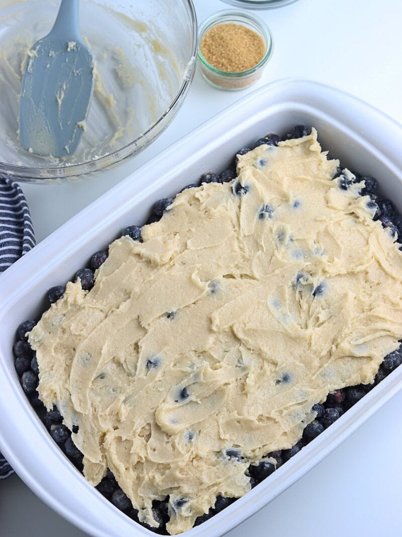 Rectangular baking dish with a layer of blueberries topped with unbaked batter; mixing bowl and spatula with leftover batter nearby.