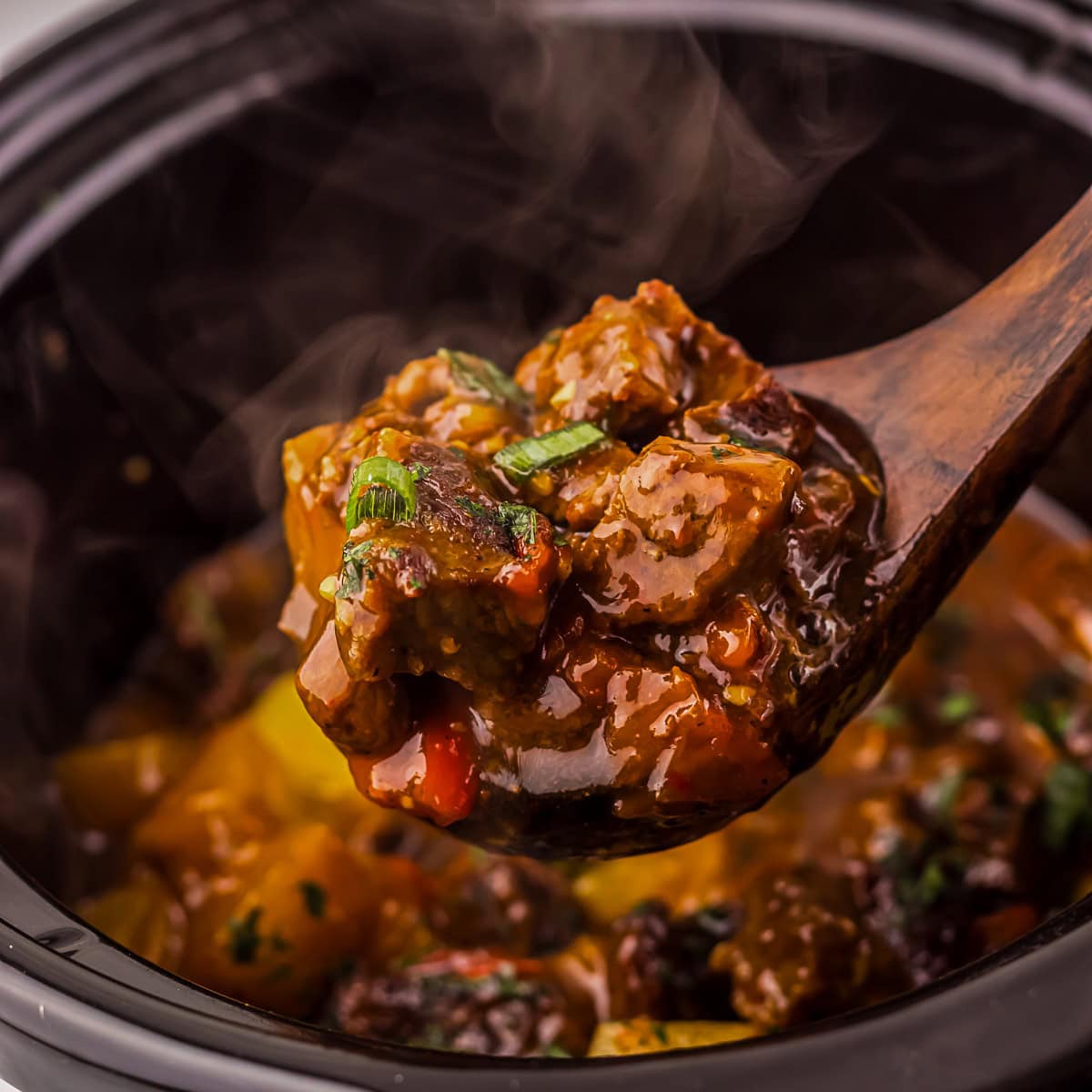 A wooden spoon lifts a serving of beef stew with vegetables and sauce from a black pot, with steam rising from the hot dish.