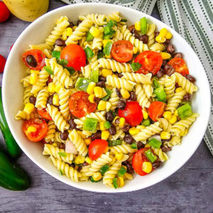 A bowl of rotini pasta salad with cherry tomatoes, black beans, corn, green bell peppers, and chopped onions on a gray surface with a striped cloth nearby.