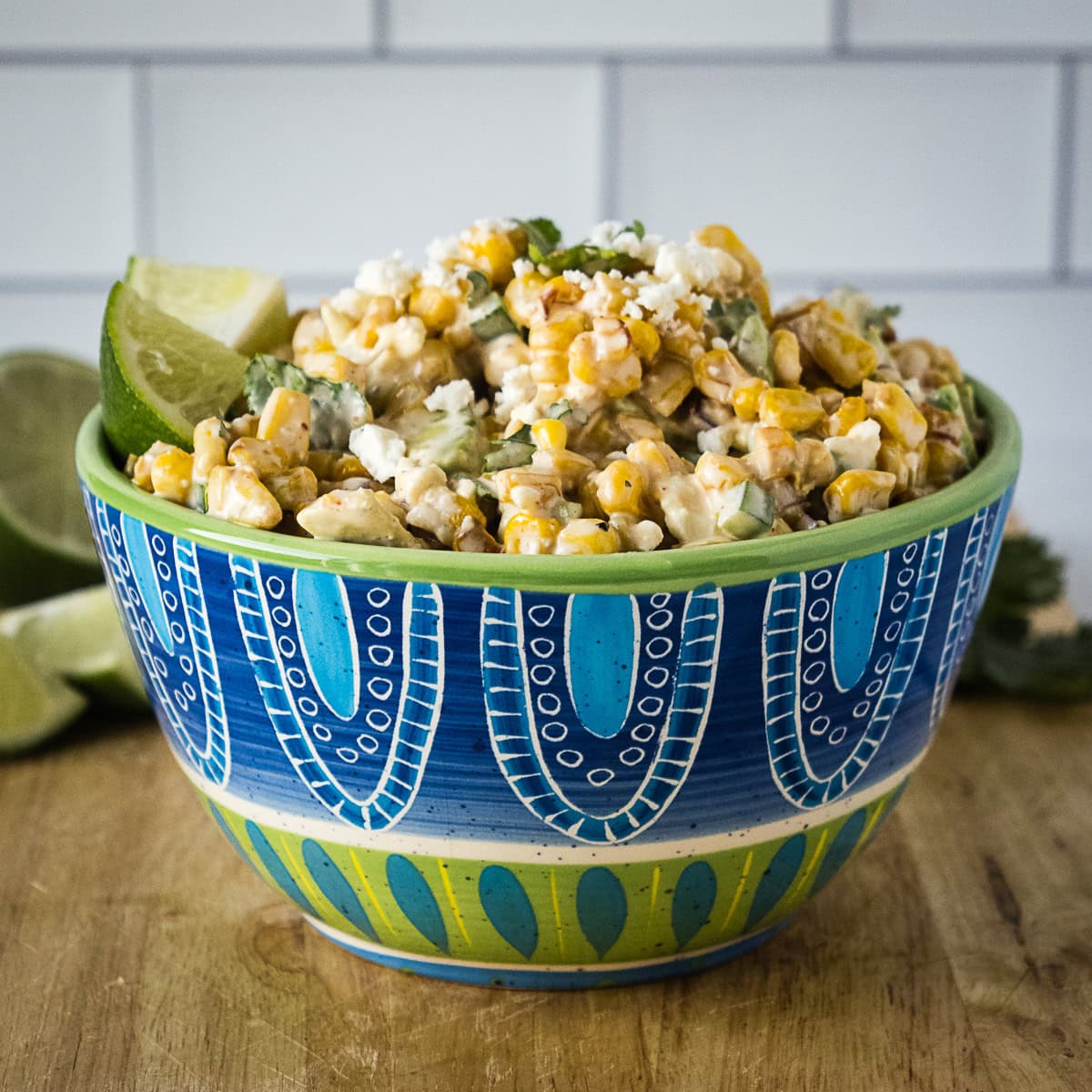 A bowl of Mexican street corn salad topped with crumbled cheese and cilantro, with lime wedges on the side, set on a wooden surface.