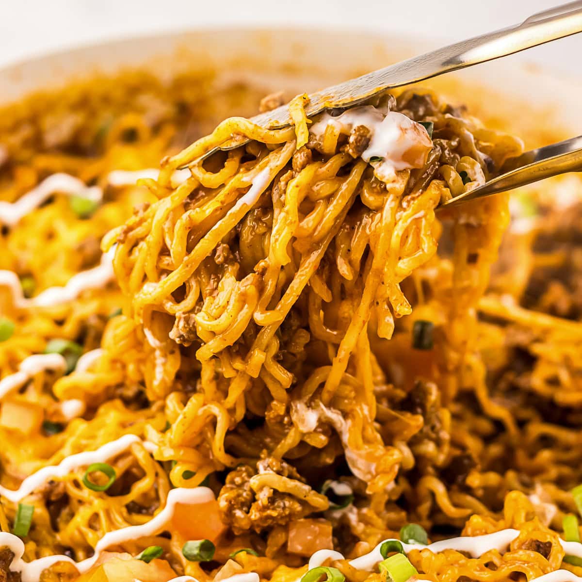 Chopsticks lifting a serving of sauced noodles with ground meat, diced vegetables, green onions, and a drizzle of white sauce.