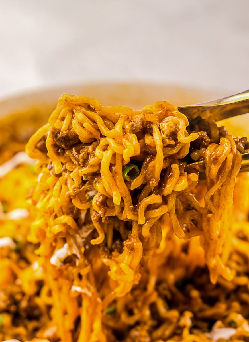 A close-up of a fork lifting a serving of cooked noodles mixed with ground beef and sauce.