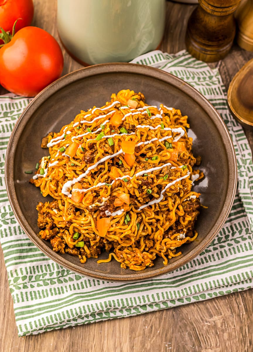 A plate of cooked instant noodles mixed with ground meat, chopped tomatoes, herbs, and drizzled with a white sauce, set on a striped cloth with tomatoes in the background.