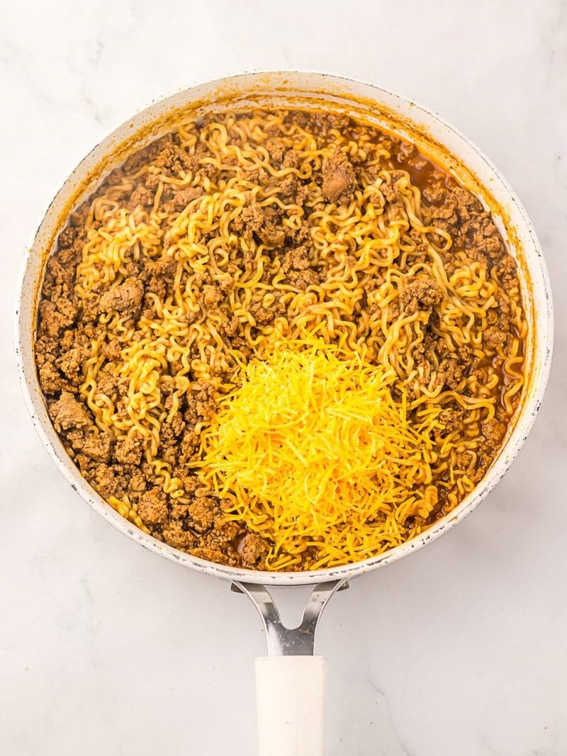 A white skillet filled with cooked ramen noodles, ground beef, and a pile of shredded cheddar cheese on top, set on a marble surface.