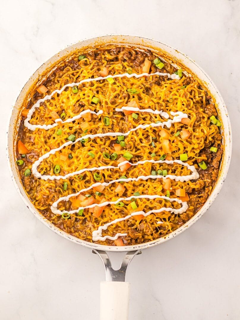 A skillet filled with cooked ramen noodles, ground meat, diced vegetables, green onions, and a drizzle of white sauce on top.