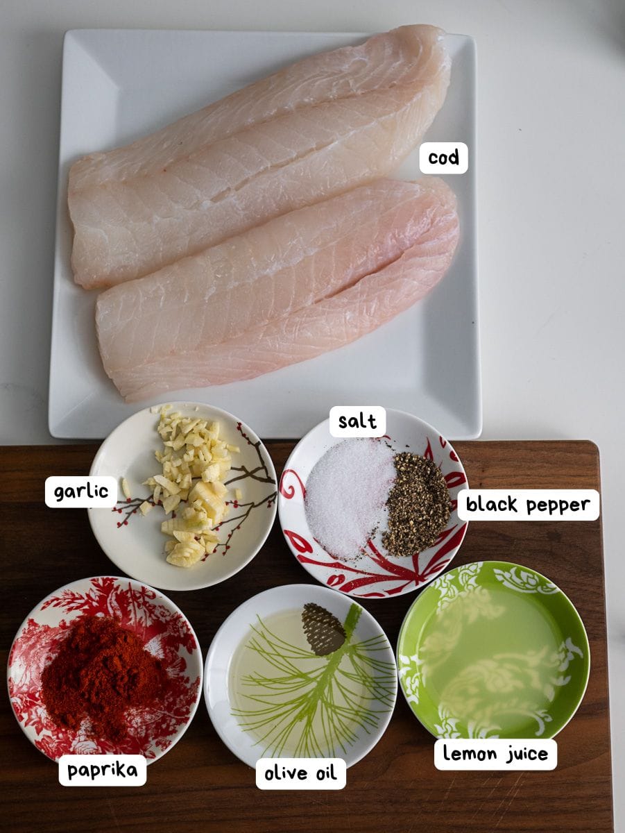 Two raw cod fillets on a plate, with small bowls of garlic, paprika, olive oil, lemon juice, salt, and black pepper arranged around them.