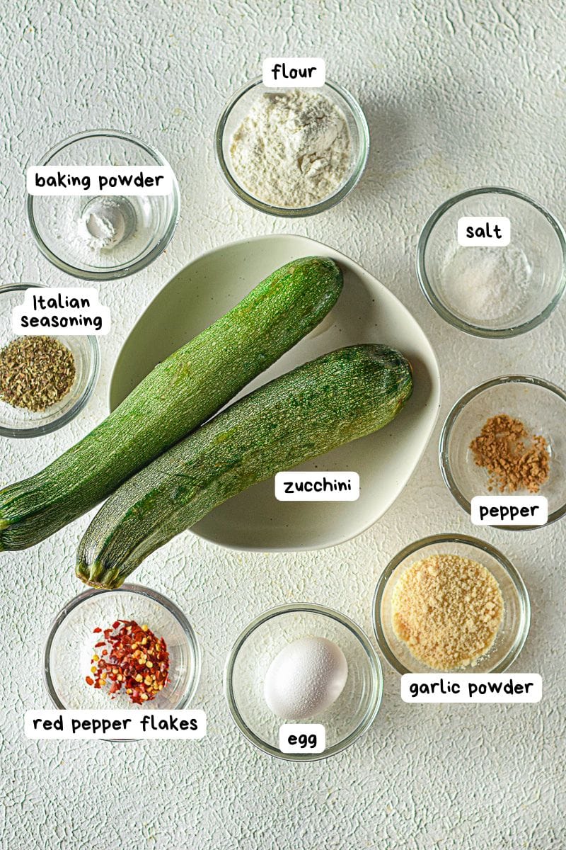 Two whole zucchinis on a plate surrounded by small bowls containing flour, salt, pepper, baking powder, Italian seasoning, red pepper flakes, garlic powder, and an egg.