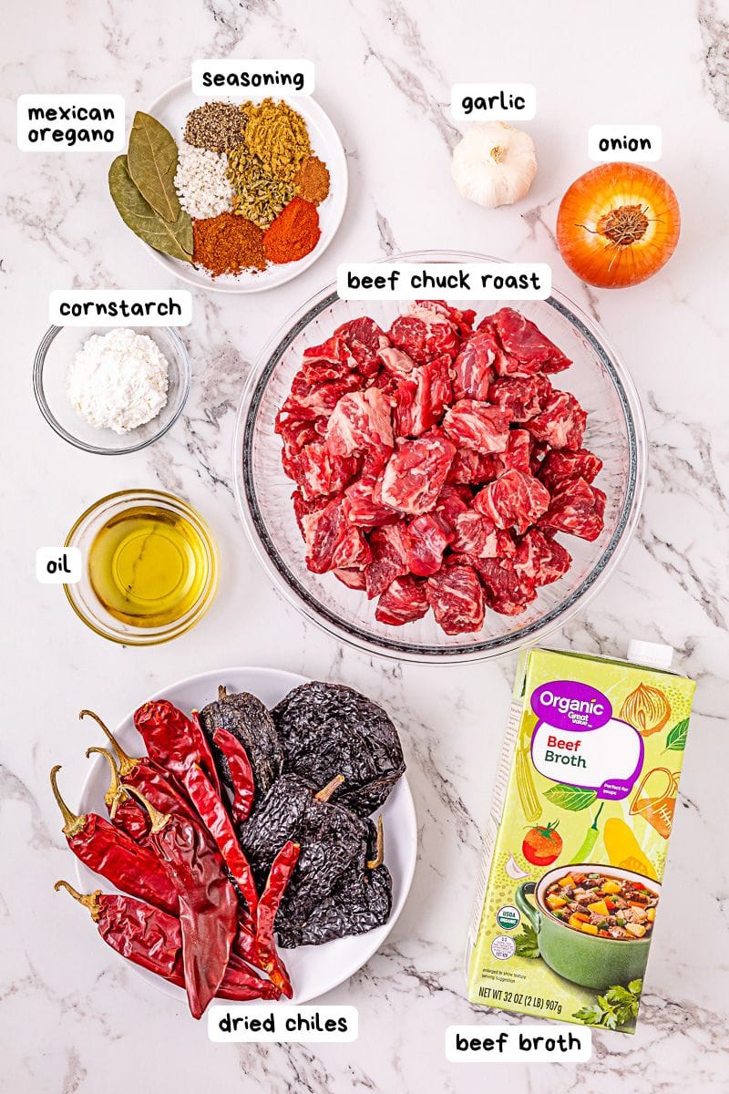Ingredients for a beef stew recipe are arranged on a marble surface, each labeled: beef chuck roast, dried chiles, oil, cornstarch, oregano, seasoning, garlic, onion, and beef broth.