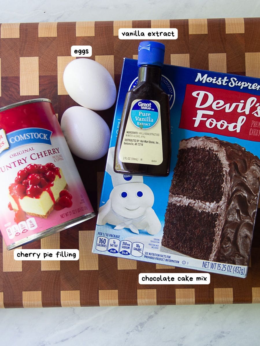 A can of cherry pie filling, two eggs, a bottle of vanilla extract, and a box of chocolate cake mix on a wooden cutting board.