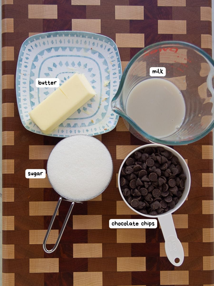 A stick of butter, a measuring cup of milk, a cup of sugar, and a cup of chocolate chips are arranged on a checkered wooden surface.
