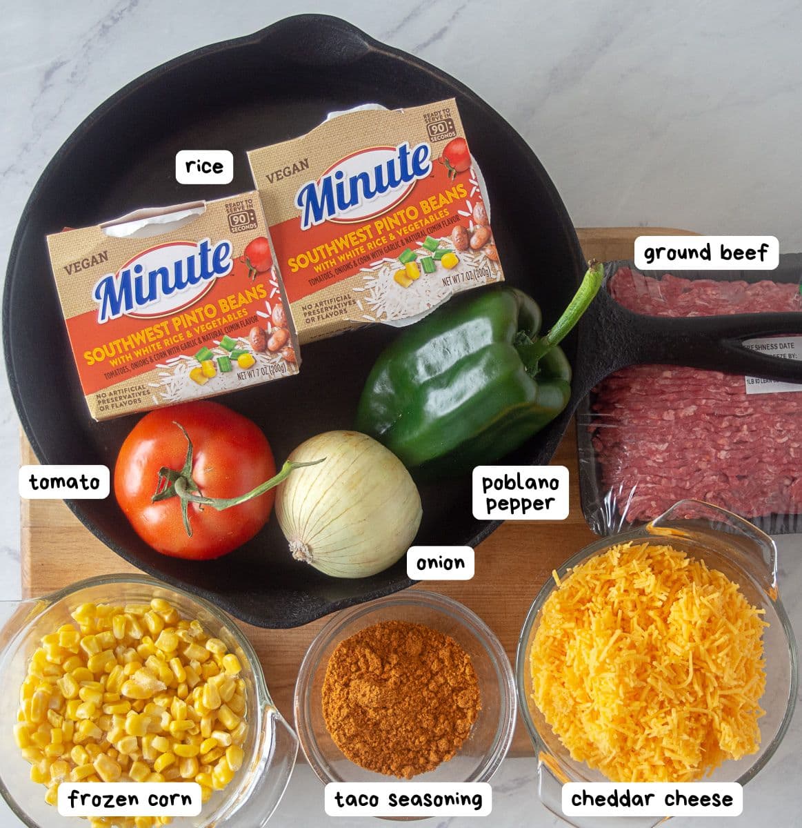 A taco rice skillet is showcased here: a cast iron skillet surrounded by packaged rice and pinto beans, poblano pepper, ground beef, tomato, frozen corn, taco seasoning, onion, and shredded cheddar cheese—all the essentials for this flavorful one-pan meal.