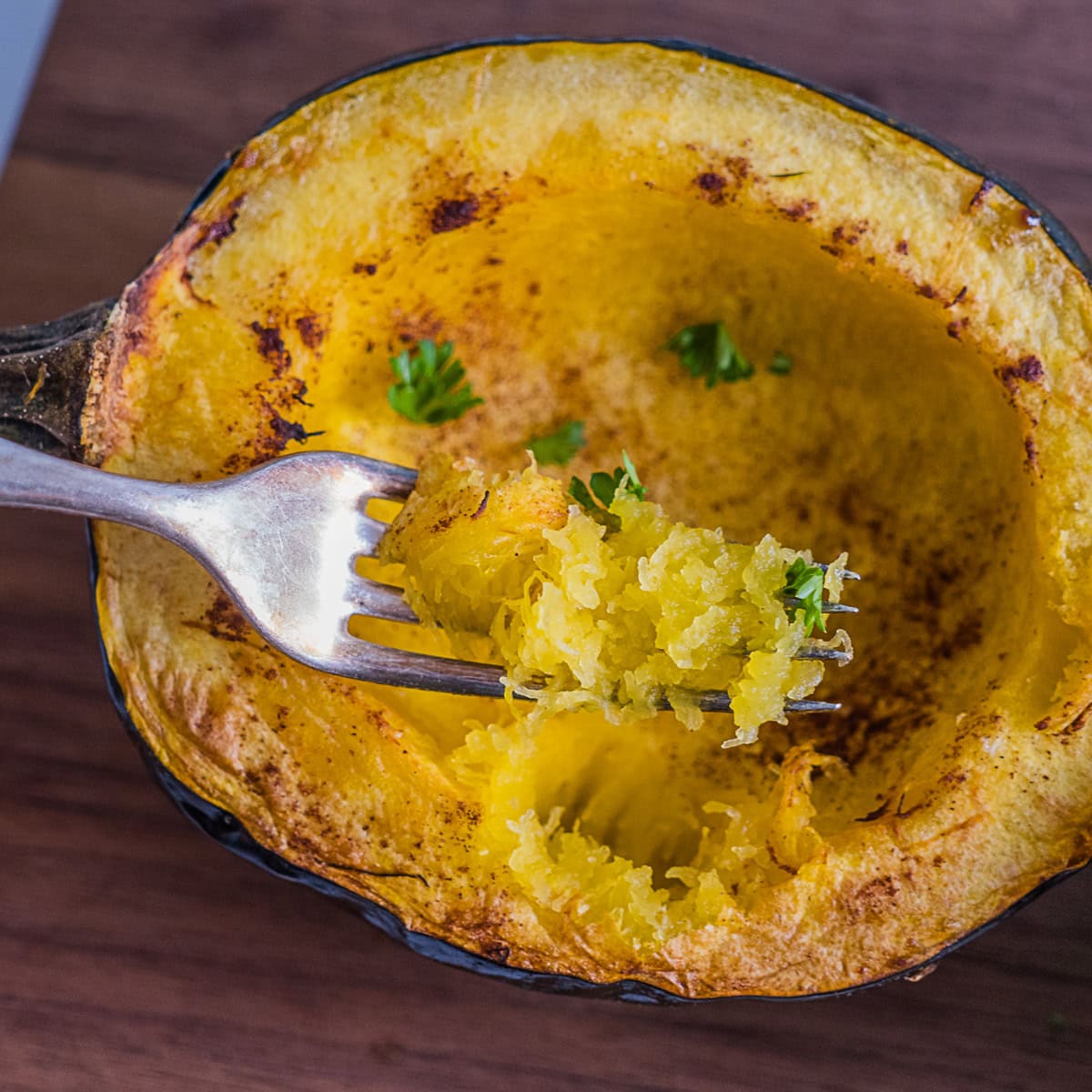 A fork holds a bite of cooked spaghetti squash inside a roasted squash half, garnished with chopped herbs, on a wooden surface.