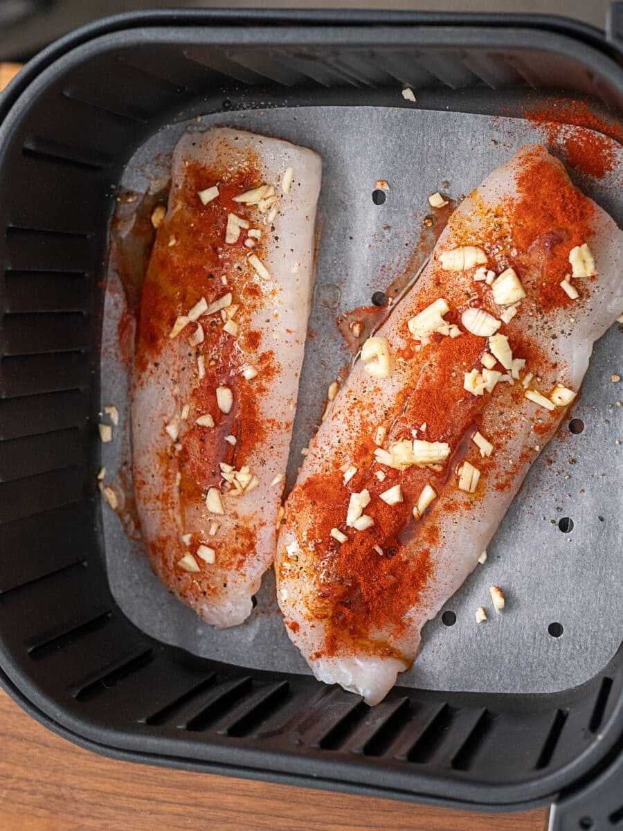 Two raw fish fillets in an air fryer basket, seasoned with paprika and chopped garlic, placed on parchment paper.