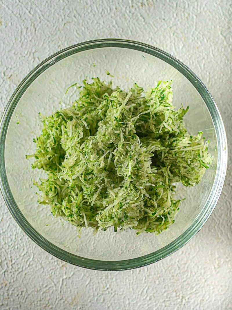 A clear glass bowl filled with shredded zucchini on a light textured surface.
