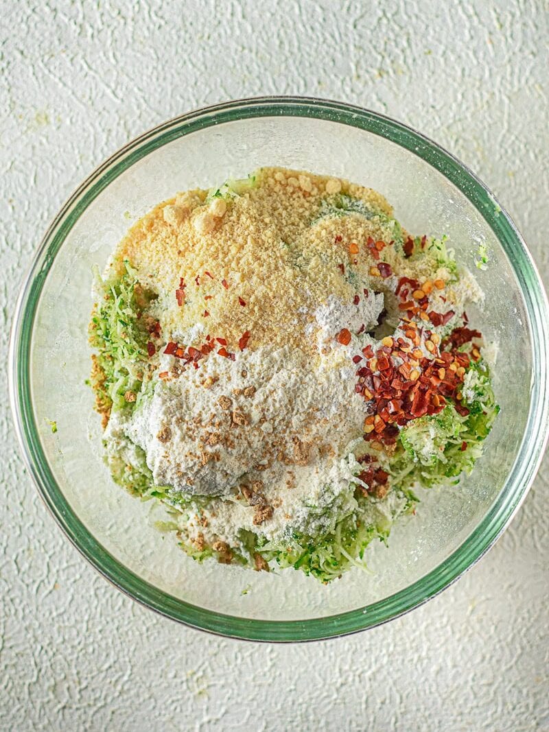 A glass bowl containing shredded zucchini, flour, breadcrumbs, red pepper flakes, and seasoning on a textured white surface.