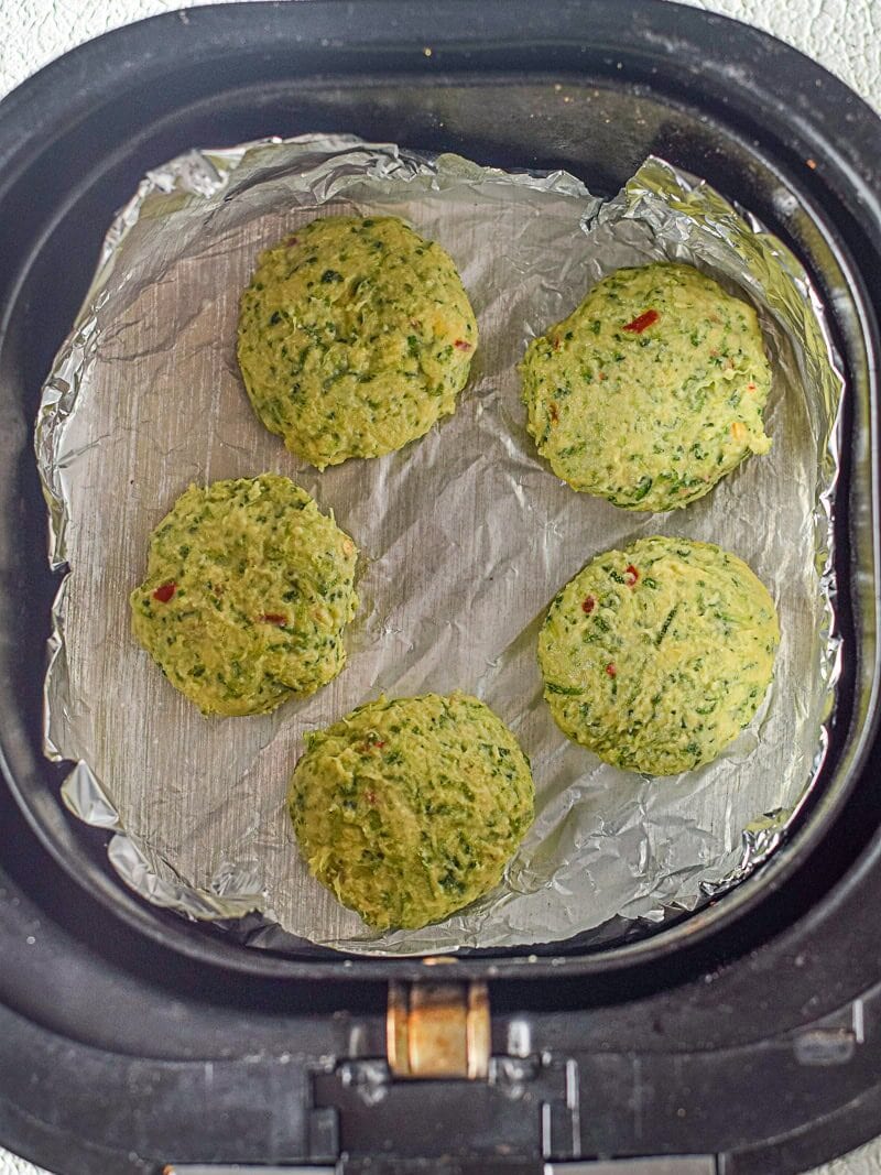 Five uncooked green veggie patties are arranged on a sheet of foil inside an air fryer basket, ready to be cooked.