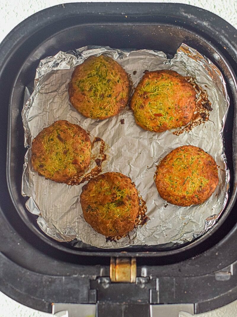 Five round, golden-brown fritters on a sheet of aluminum foil inside an air fryer basket.