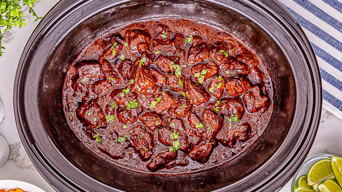 Chunks of beef in a rich, dark sauce garnished with chopped parsley, served in a black slow cooker on a white countertop.